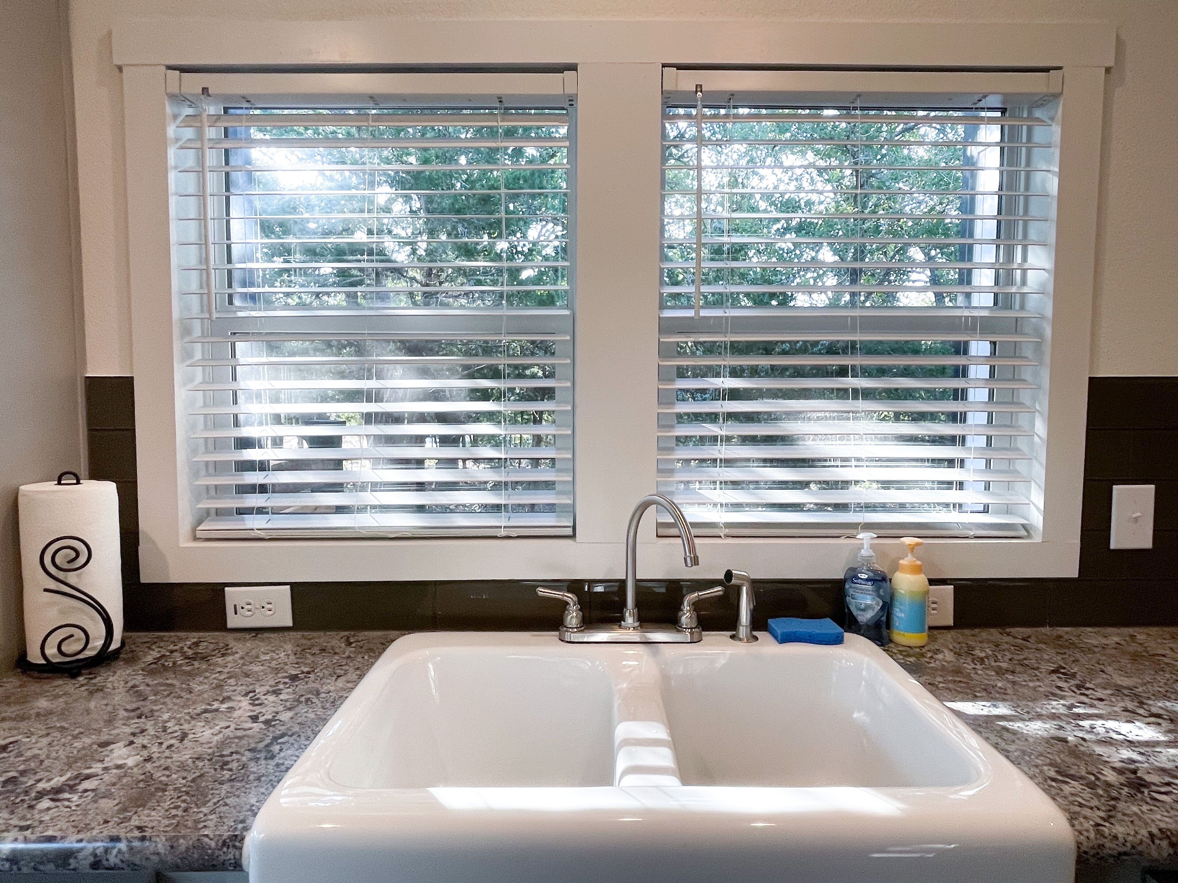A bathroom with a white sink, two windows with blinds, a towel dispenser, and a soap dispenser.