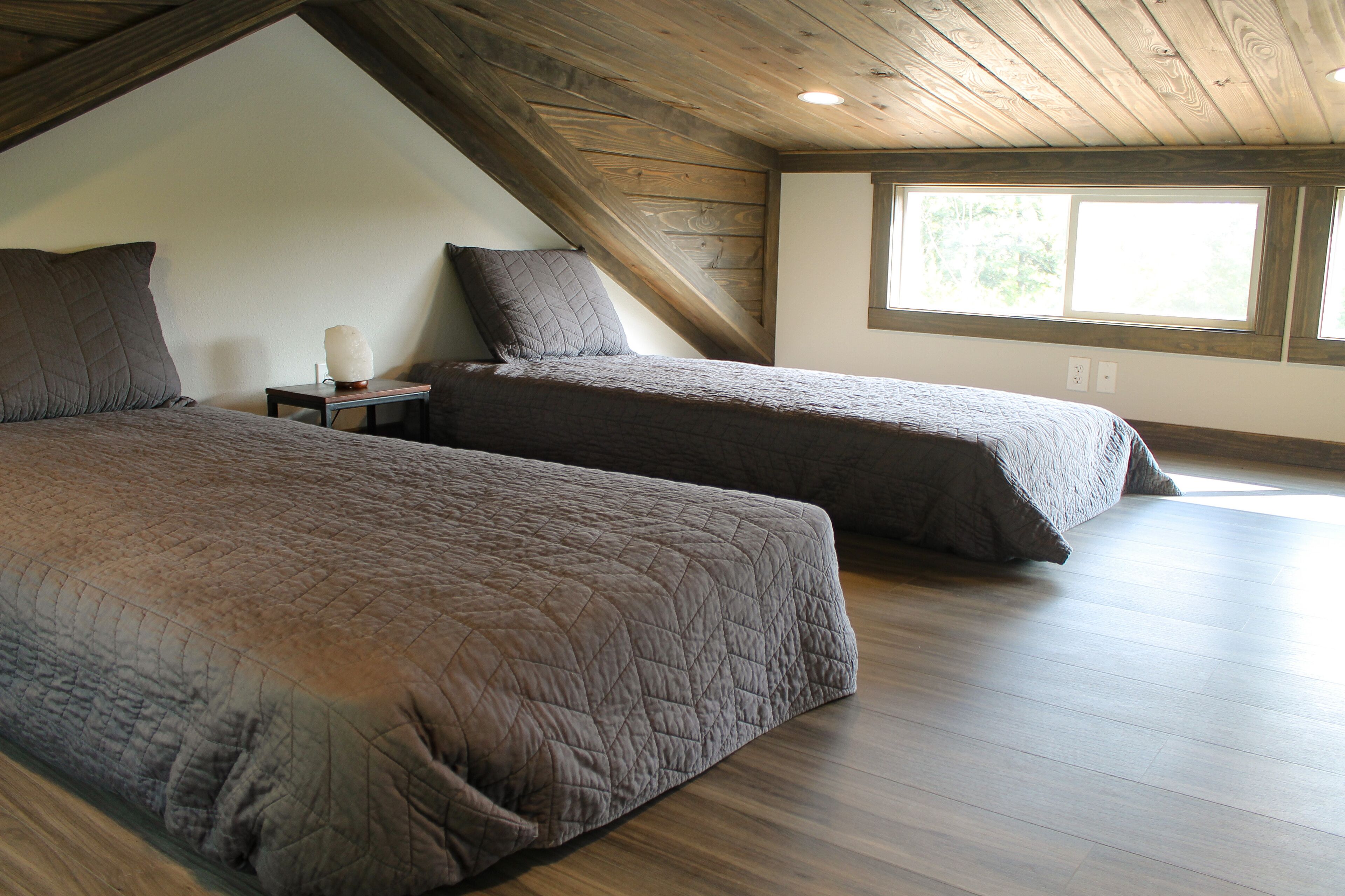 Une chambre avec deux lits, un plancher en bois, un plafond en pente et une fenêtre laissant entrer la lumière naturelle.