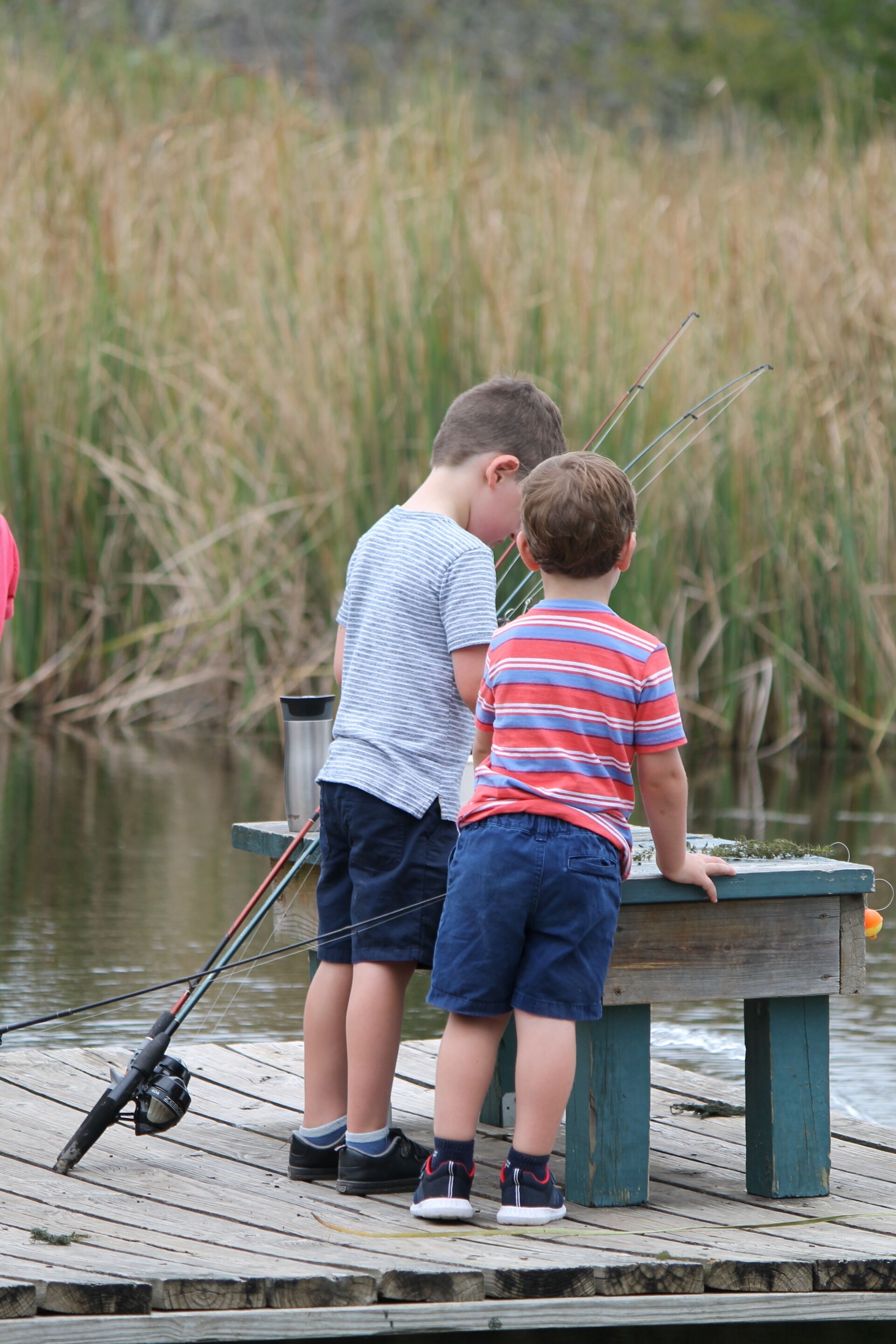 Two children fishing on a wooden dock with fishing rods and a metal container.