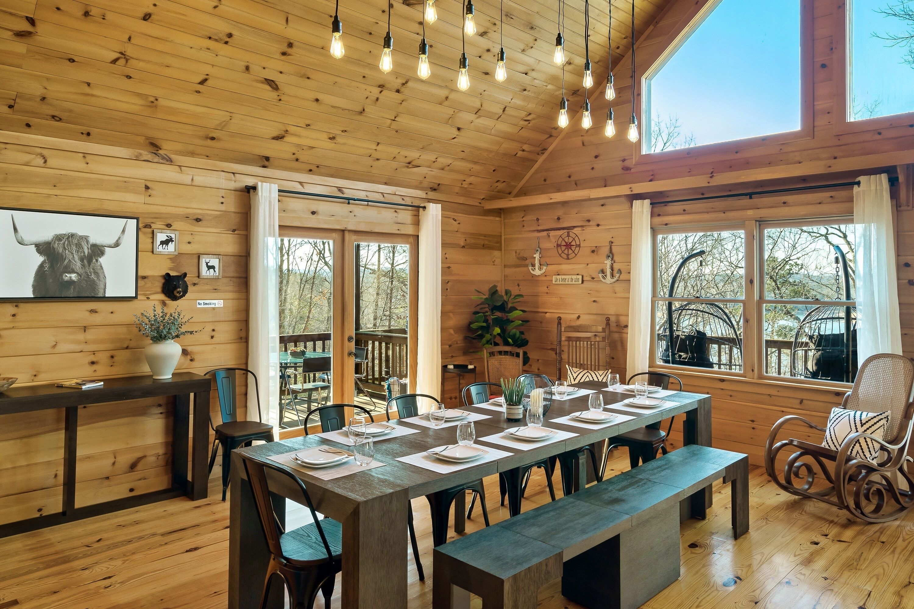 A wooden cabin interior with a dining table set for four, a bench, and a view of the outdoors through large windows.