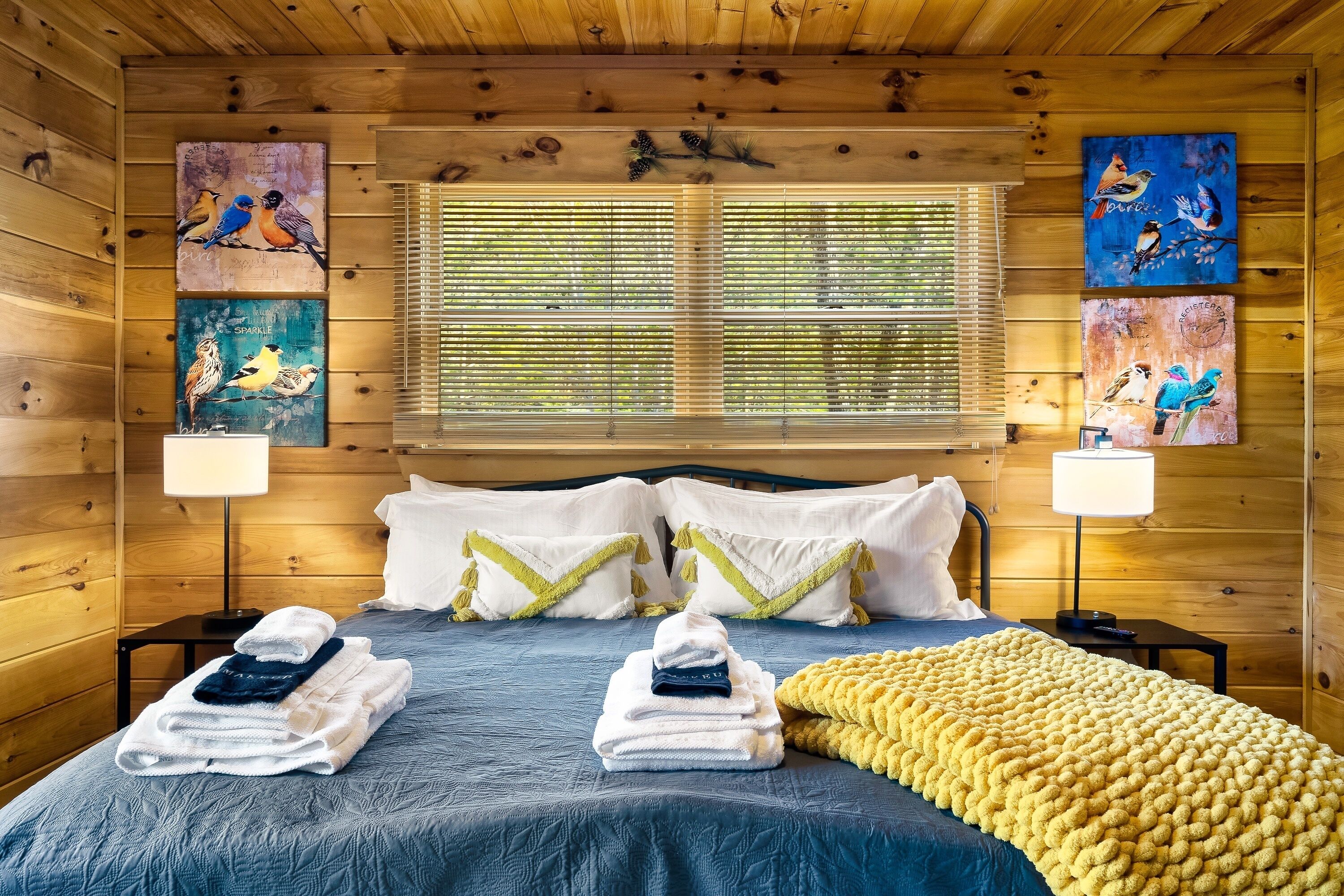 A wooden cabin room with a bed, two wall-mounted lamps, and three bird-themed paintings.