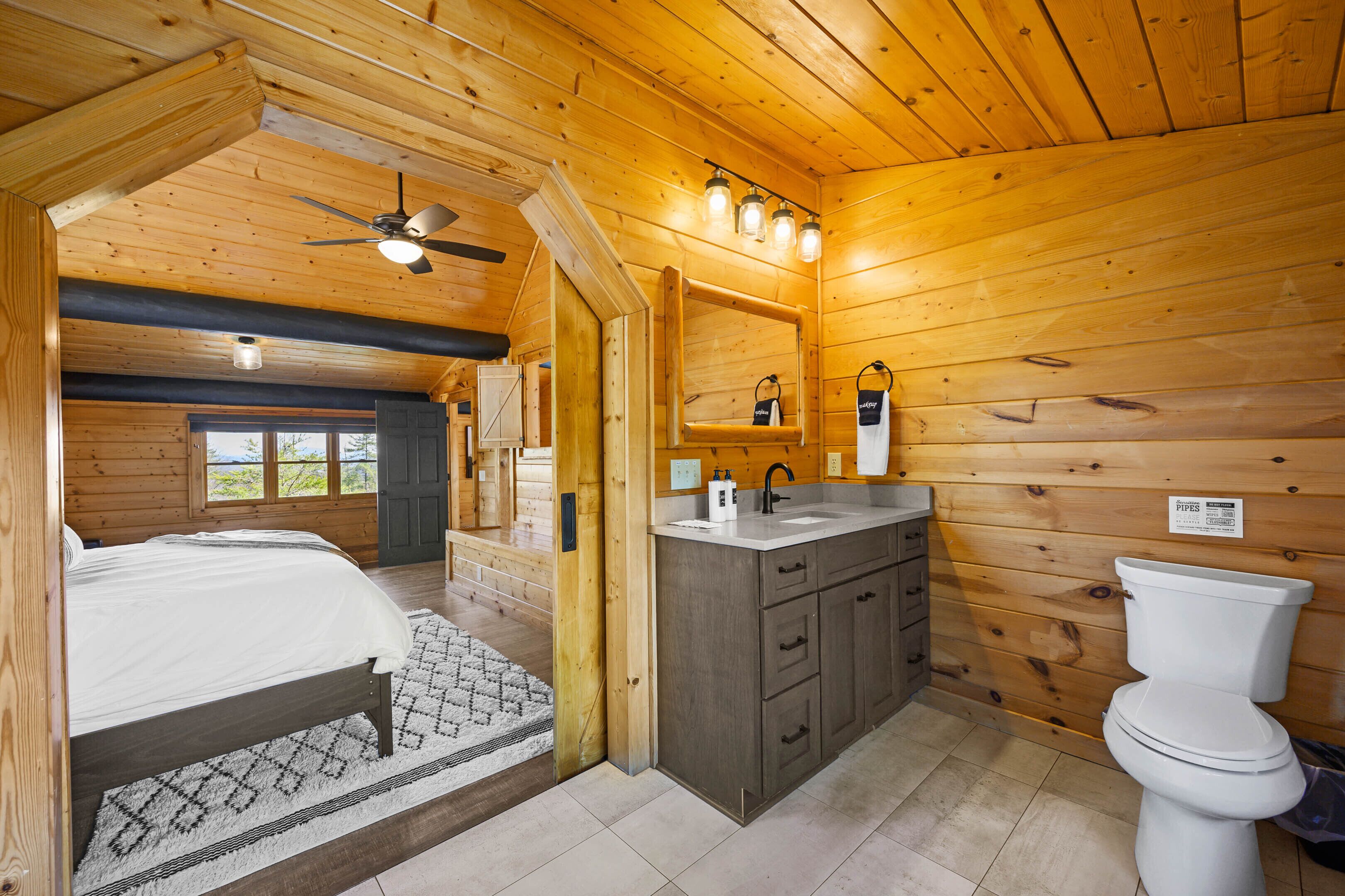 A wooden cabin interior with a bed, a bathroom with a sink and toilet, and a ceiling fan.