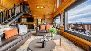A log cabin interior with a staircase, leather sofa, coffee table, and a large window offering a mountain view. - STUnning Views|Hot tub|FirePit|Easy Drive|Pool Table|Semi Secluded|Easy CheckOut (Sevierville)