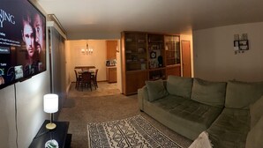 Un soggiorno con un divano verde, una TV montata a parete, un'area pranzo illuminata da un lampadario e un mobile contenitore in legno con vetrate.