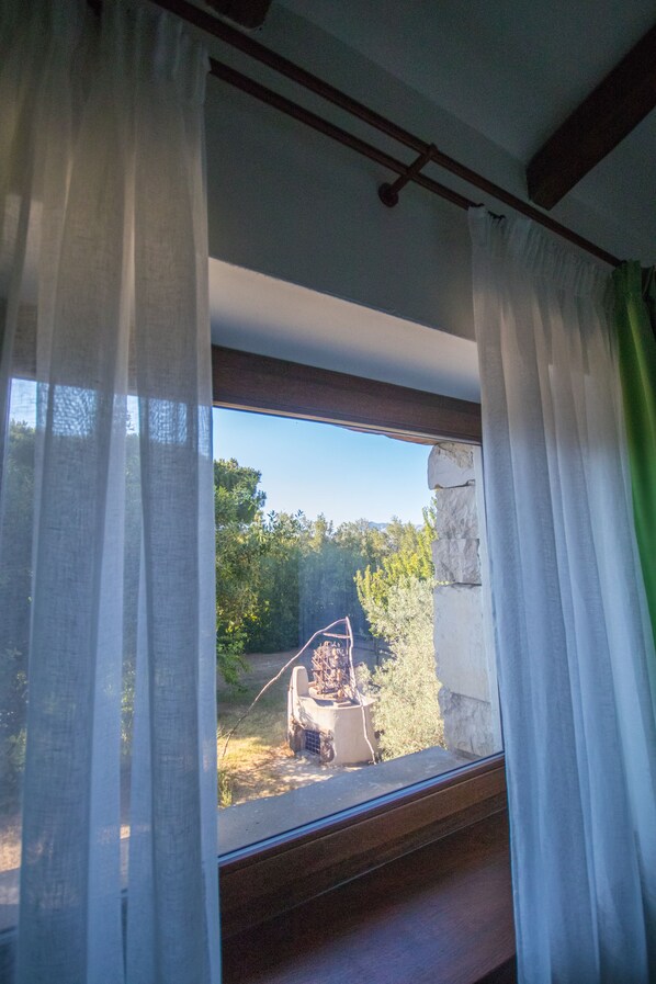 Una ventana con cortinas traslúcidas, vista a un jardín y un muro de piedra.