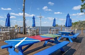 Eine Holzterrasse mit Picknicktischen und blauen Sonnenschirmen, mit Blick auf eine Marina und Boote.