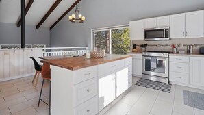A modern kitchen with white cabinets, a wooden island, stainless steel appliances, and a large window offering a view of trees. - Pine Knoll - Peaceful, Hot Tub, Wifi, Propane Fire Pit, Newly Furnished (Bend)