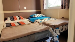 Une chambre de taille modeste, comprenant deux lits, une fenêtre avec des rideaux, une étagère ornée d’objets décoratifs et une petite plante.