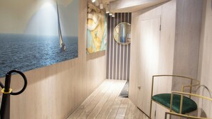 A hallway with a wooden floor, a green chair, a wall-mounted mirror, and a painting of a sailboat. - Hotel Velero (Rionegro)