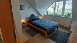 Ein Schlafzimmer mit schrägen Wänden, einem Bett mit blauer Bettdecke, einem kleinen Tisch mit Lampe und einem Fenster mit Blick auf Bäume.