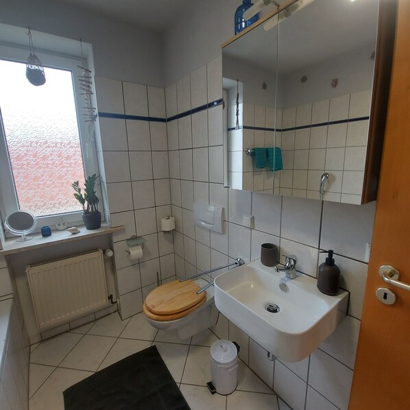 A small, clean bathroom with a toilet, sink, and mirror. There's a window with a view, a radiator, and a small shelf with toiletries. - Edder Apartment - a quiet and cozy vacation apartment with a large balcony (Edermünde)