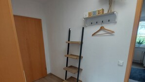Ein Zimmer mit Holzboden, einem schwarzen Metall-Leiterregal, einer Holztür, einer weißen Wand mit dem Schriftzug 'HOME' und einem hängenden Holzaufhänger.