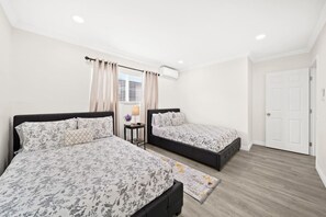 A bedroom with two beds, a window with curtains, an air conditioning unit, and a door.