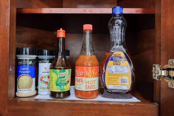 Un mueble con cuatro botellas de condimentos: ajo en polvo, sazonador líquido, salsa picante y sirope para panqueques con sabor a mantequilla.