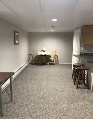 A small, carpeted room with a round table and four chairs, a small stool, and a kitchenette with wooden cabinets. - The Cabin @Lake George (Lake George)