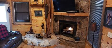 Ein gemütliches Blockhäuschen im Inneren mit Kamin aus Stein, Holzwänden, einer Ledersofa und einem Wand-Fernseher.