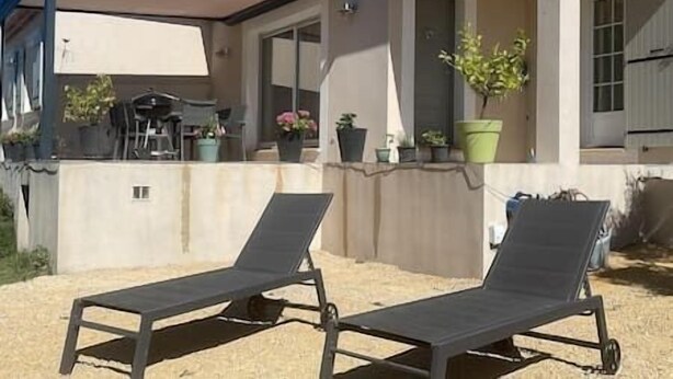 A covered outdoor seating area with two lounge chairs, a patio table, and potted plants. - To complete (Aramon)