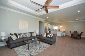 A modern living room with a gray sectional sofa, a coffee table, and a dining area with a table and chairs. A ceiling fan and recessed lighting are visible.