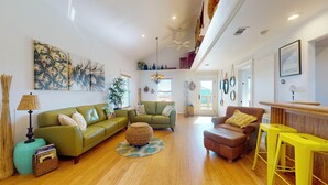 Ein geräumiges Wohnzimmer mit einem grünen Sofa, einem braunen Sessel und einem gelben Tisch. Es gibt ein großes Fenster mit Jalousien, einen Deckenventilator und eine Wanduhr.