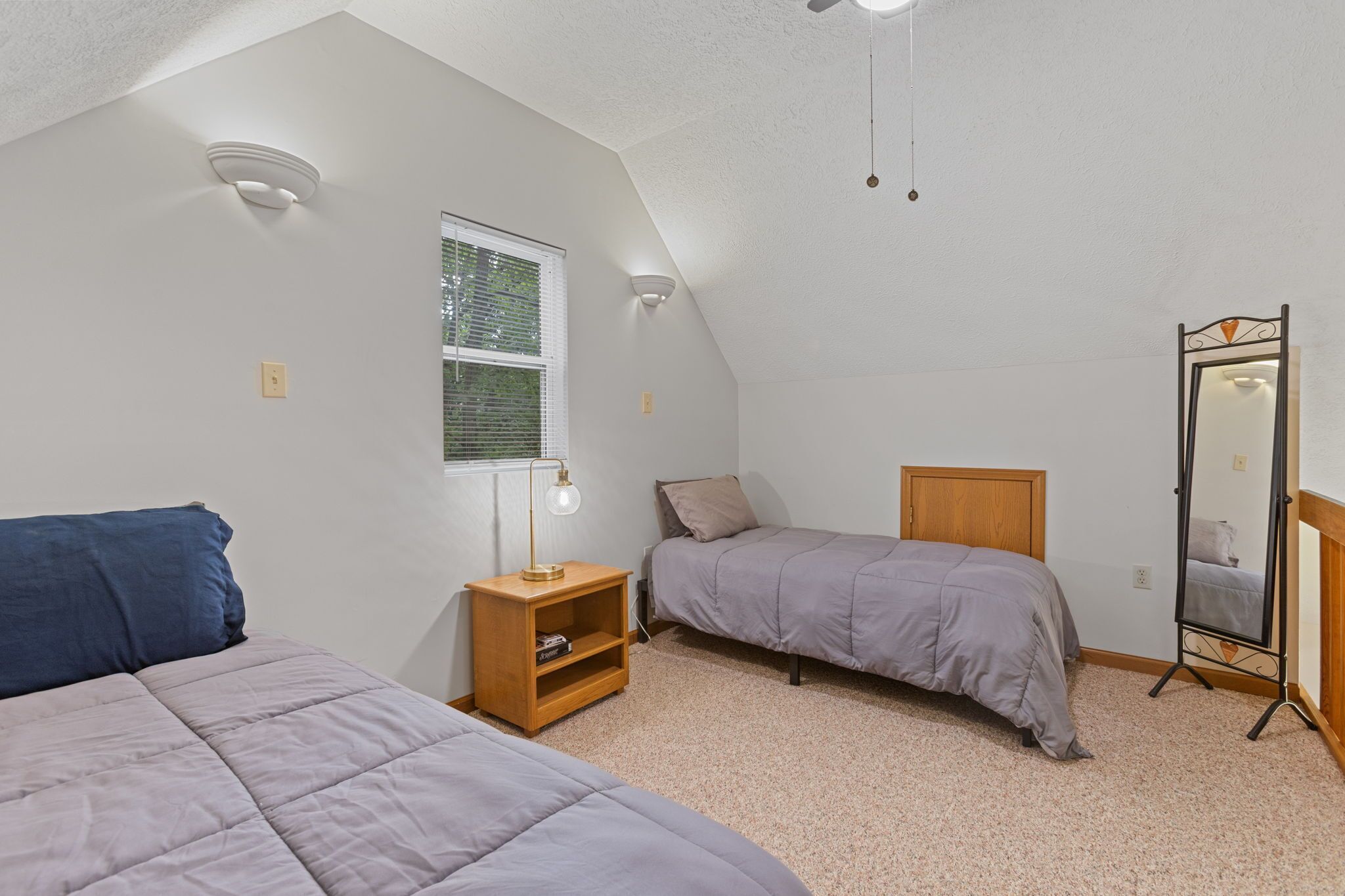 A bedroom with two single beds, a nightstand with a lamp, a mirror, and a window with blinds.