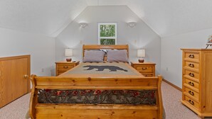 Ein Schlafzimmer mit schrägem Dach, einem Bett mit Bärenmotiv, zwei Nachttischen mit Lampen, einer Kommode und einem Fenster mit Blick auf Bäume.