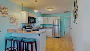 Una cocina con armarios blancos, microondas, estufa y refrigerador. Hay dos taburetes de bar y una ventana con persianas.