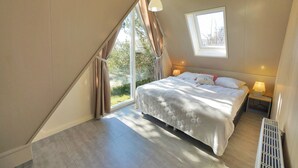 Een slaapkamer met een schuine plafond, een bed met een witte dekbedovertrek en kussens, een nachtkastje met een lamp, en een raam met gordijnen.