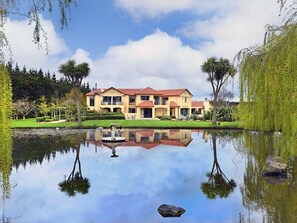 A large, two-story house with a red roof, surrounded by a well-maintained garden and a pond reflecting the building. - Rural luxury - 5 bedroom getaway (Kirwee)