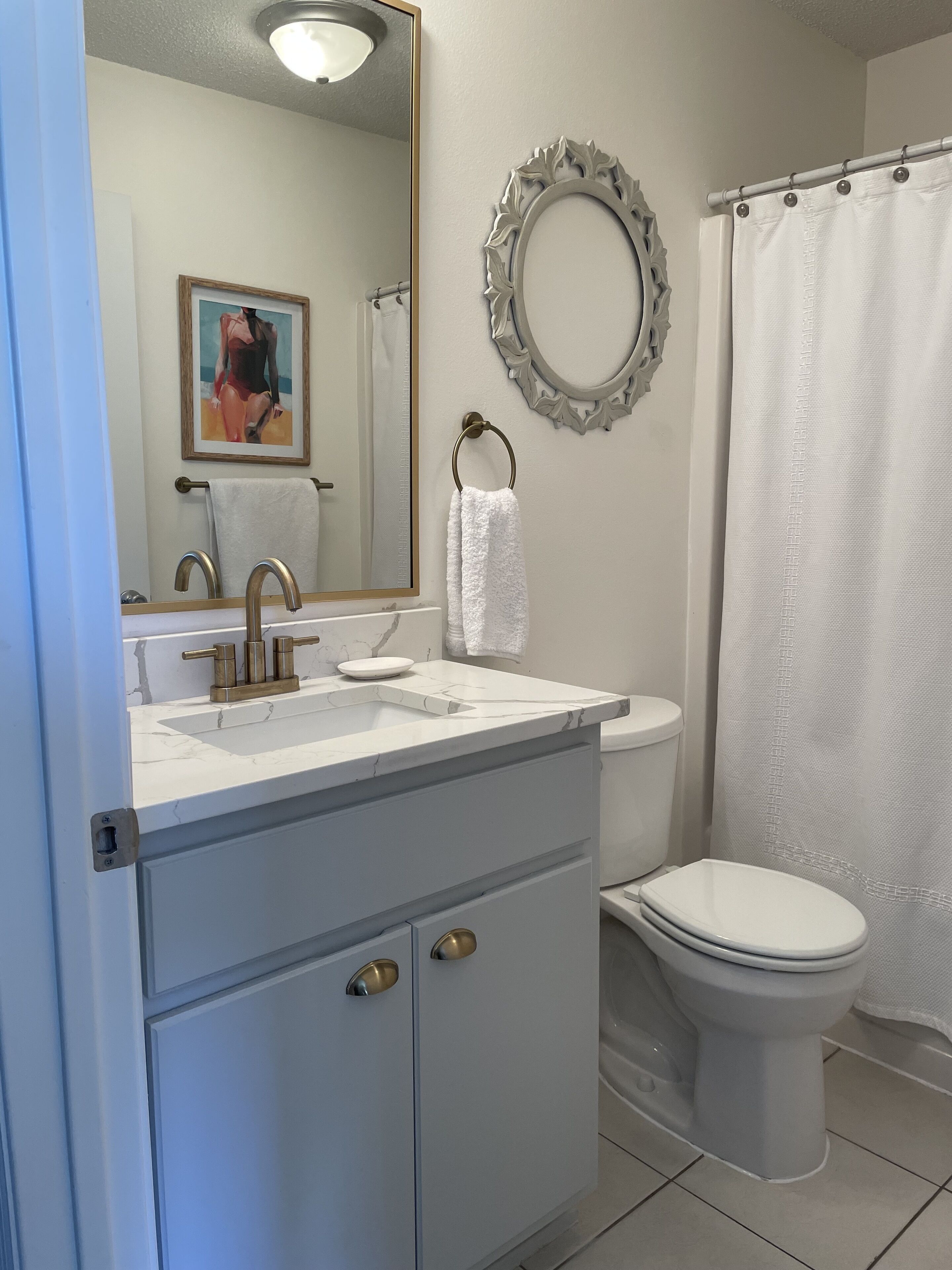 Une salle de bain avec un lavabo blanc, un miroir et des toilettes. On y trouve un porte-serviettes avec une serviette blanche et un tableau encadré accroché au mur.