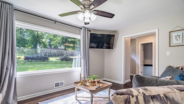 A living room with a ceiling fan, a TV mounted on the wall, a wooden coffee table with decorative items, a gray sofa, and a view of the outdoors through a large window. - Marietta! Near the Square, Pets & Long stays ok. Entire home (Marietta)