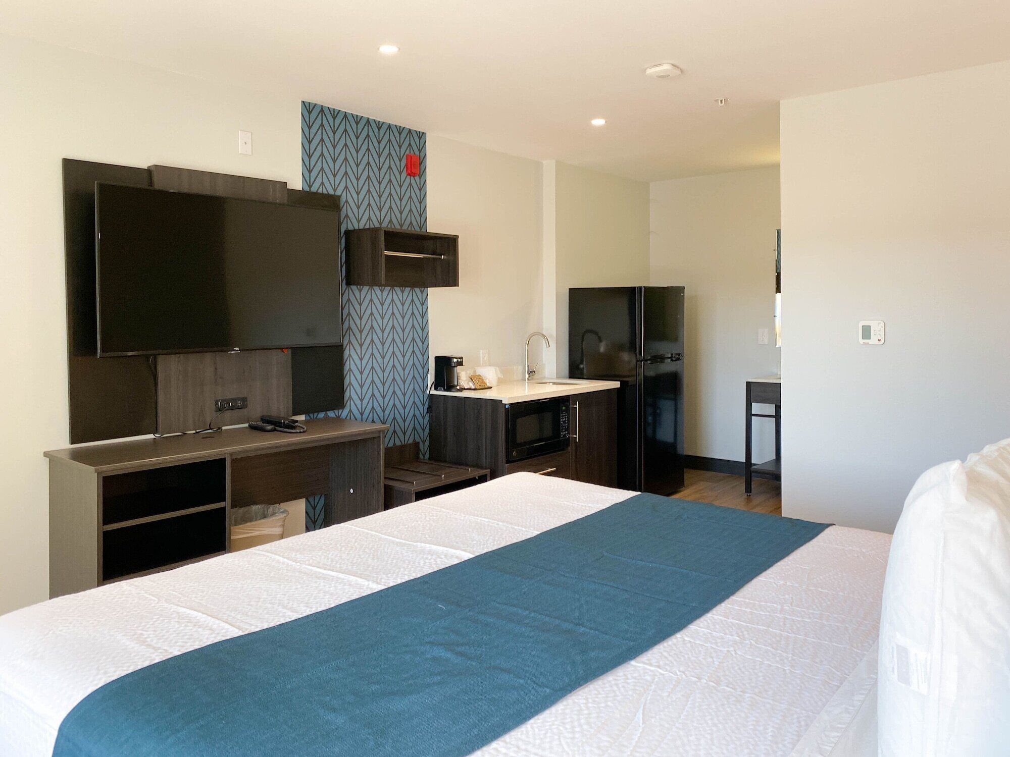 A hotel room with a bed, a TV, a desk with a chair, a microwave, a refrigerator, and a small kitchenette.