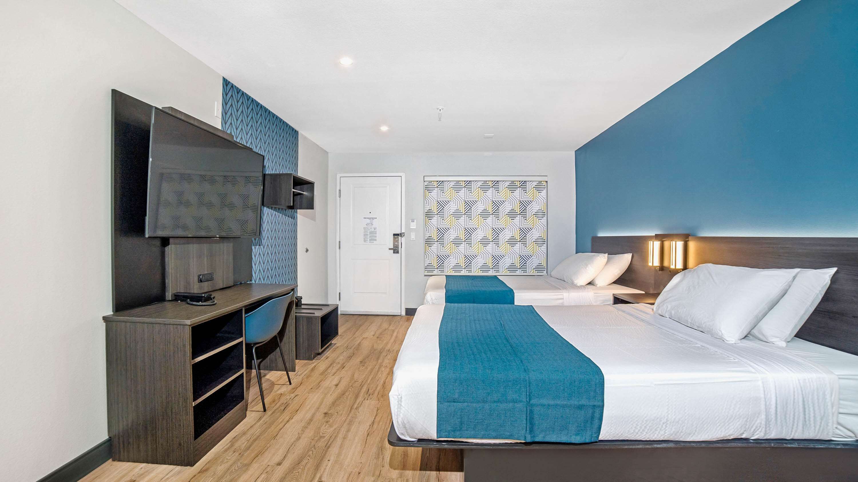 A modern hotel room with a large bed, a desk with a chair, a TV, and a patterned wall.