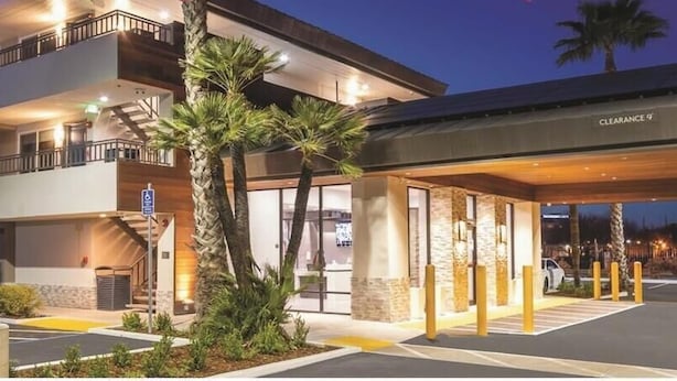A multi-story hotel with a covered entrance, a parking lot, and palm trees. - Studio 6 Suites – Stockton, CA – Waterfront (Stockton)