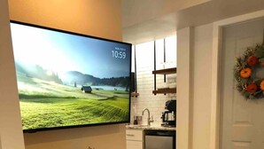Un salon moderne avec un téléviseur à écran plat fixé au mur, une cheminée, une table basse en bois et un coin cuisine à l’arrière-plan.
