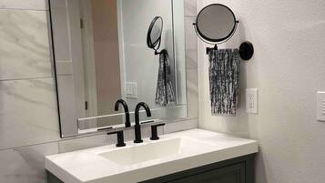 A bathroom with a white sink, two black faucets, a large mirror, a towel rack with a patterned towel, and a toilet with a roll of toilet paper.