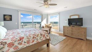 A bedroom with a bed, a television, a ceiling fan, a dresser, and a view of the ocean through large windows. - Dolphin Watch - 7Br - Oceanfront - Pool/Hot Tub (Kure Beach)