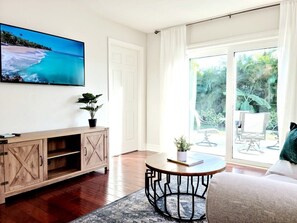 Ein Wohnzimmer mit Holzschrank, rundem Tisch, einer Couch und einem großen Fenster mit Blick auf Grünflächen.