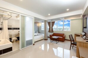 A modern hotel room with a bathroom, a bed, a sofa, a desk, and a view of the city.