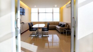 Una sala de conferencias moderna con un separador de vidrio, una mesa blanca con cuatro sillas, un sofá y un televisor montado en la pared.