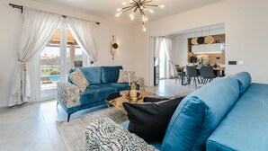 Una sala moderna con un sofá azul, una mesa de centro y una araña. Se ve un comedor con una mesa y sillas a través de una puerta abierta.