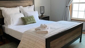 Un lit bien fait, avec du linge de lit blanc et une couverture à rayures, une table de chevet avec une lampe, et une vue sur l’extérieur par une fenêtre ornée de rideaux.
