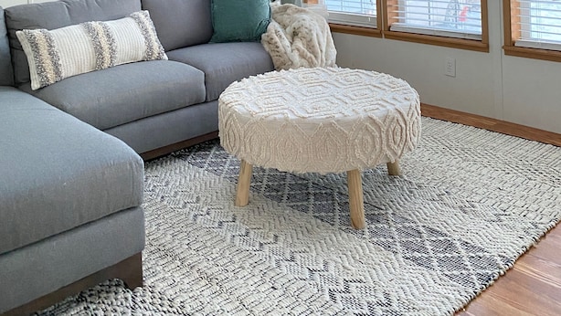 Un salon avec un canapé d’angle gris, un pouf rond, un ventilateur de plafond et de grandes fenêtres équipées de stores.