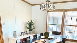 Un coin repas avec une table en bois, des chaises, un lustre, une plante en pot et une fenêtre avec des stores.