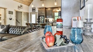 Um interior rústico com teto de madeira, sofá de couro, mesa de jantar com frutas e uma garrafa, e uma cozinha ao fundo.