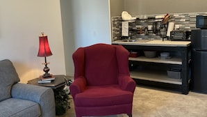 Ein Wohnzimmer mit einem roten Sessel, einem grauen Sofa, einem kleinen Tisch mit Lampe sowie einer Küchenzeile mit Mikrowelle und Toaster.