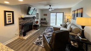 Una sala de estar moderna con chimenea, un televisor de pantalla plana montado encima, un sofá, una mesa de centro y vistas a un balcón exterior a través de una puerta corrediza de vidrio.