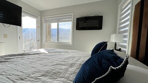 Un dormitorio con una cama grande, un televisor montado en la pared, una ventana con vistas a las montañas y un balcón.