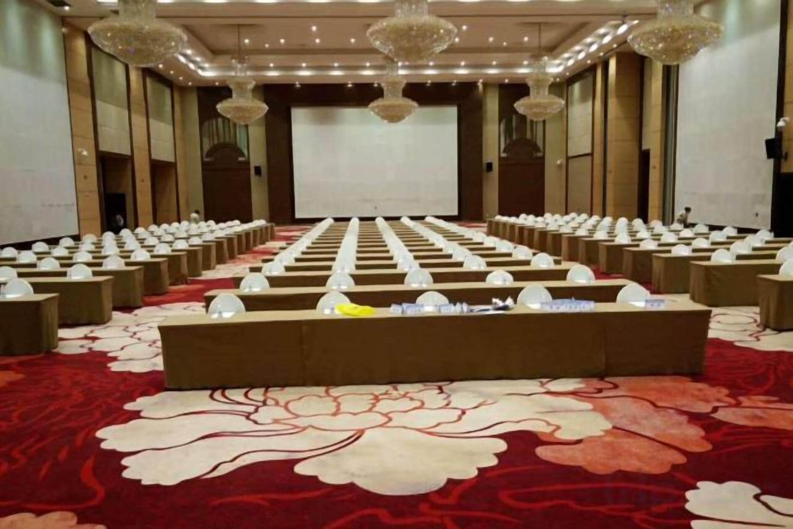 A conference room with rows of tables and chairs, a large screen at the front, and a red carpeted floor.