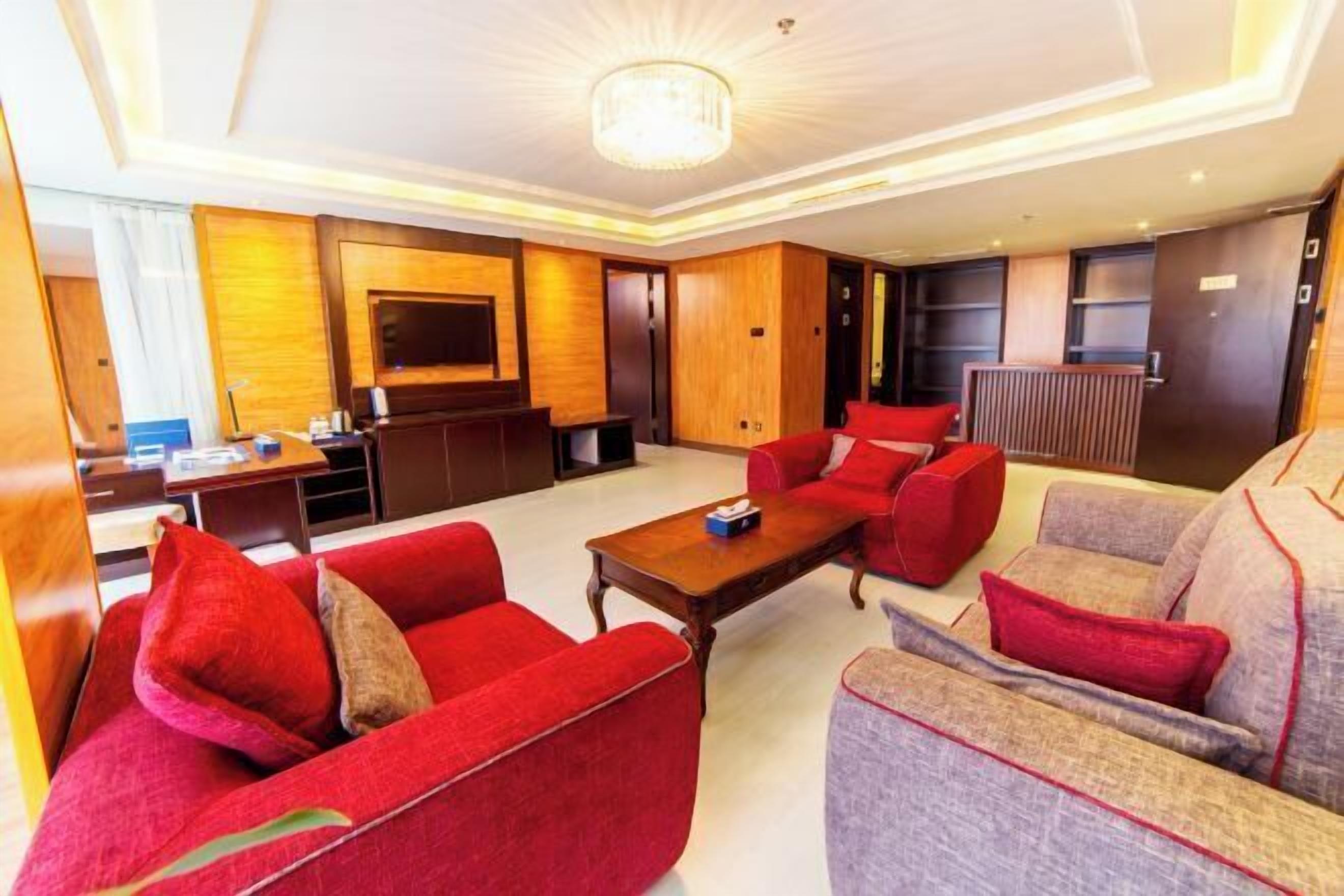 A hotel room with a large flat-screen TV, a wooden accent wall, a red sofa, a grey sofa, and a wooden coffee table.