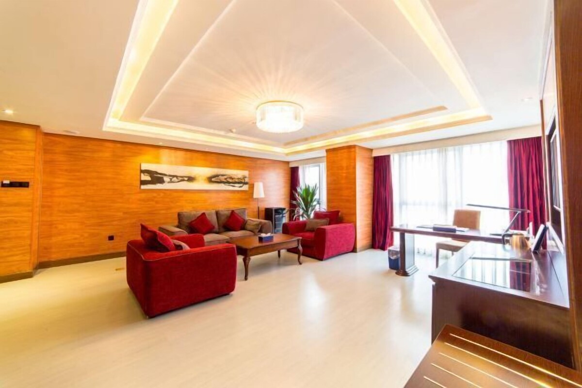A modern hotel room with a wooden accent wall, a red sofa set, a dining area with a glass table, and a large window with curtains.